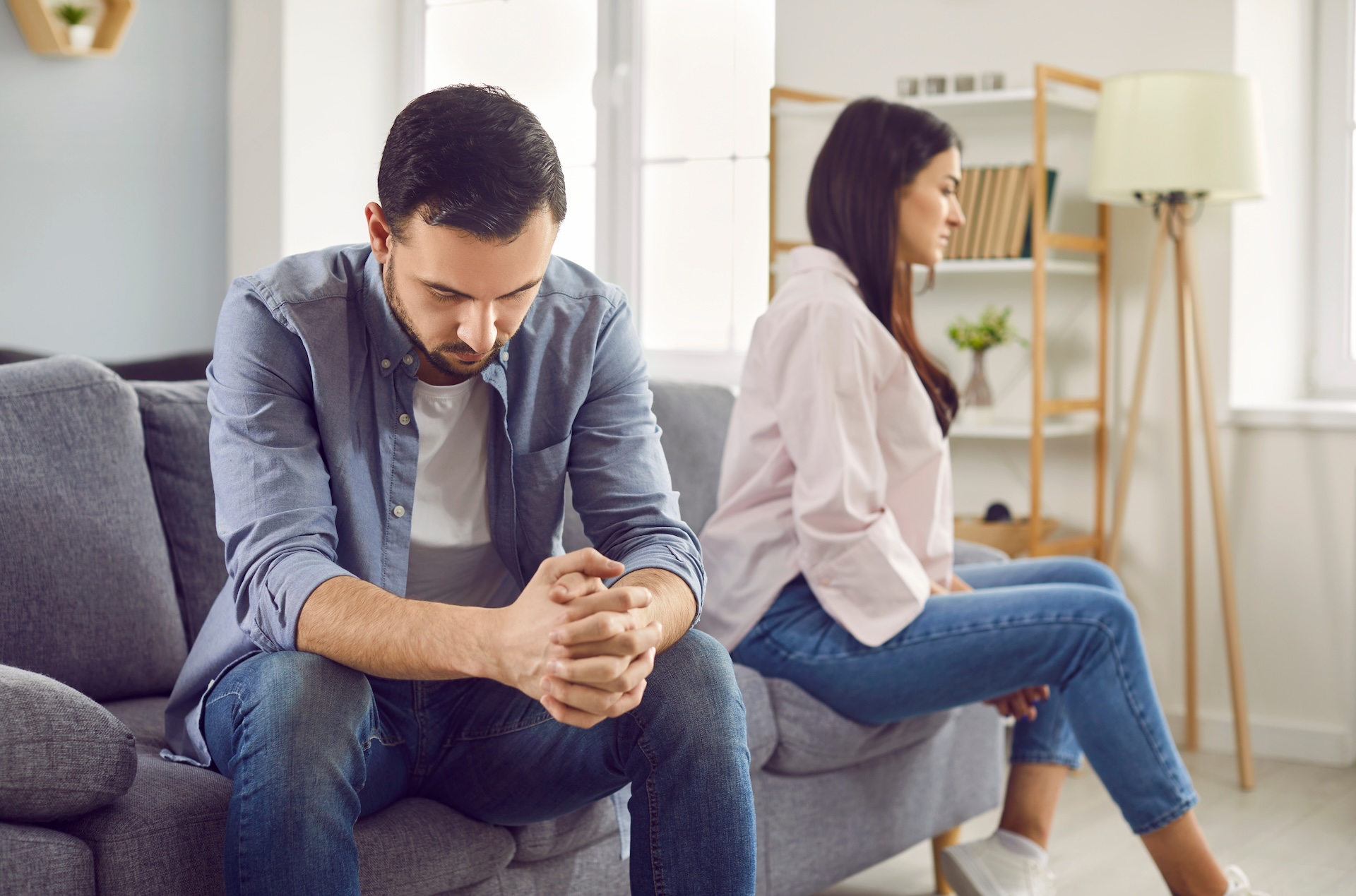 Upset young man sitting on the sofa and a woman in background ignoring each other at home. Stressed married couple sitting separately in the living room. Upset young man sitting on the sofa and a woman in background ignoring each other at home. Stressed married couple sitting separately in the living room.