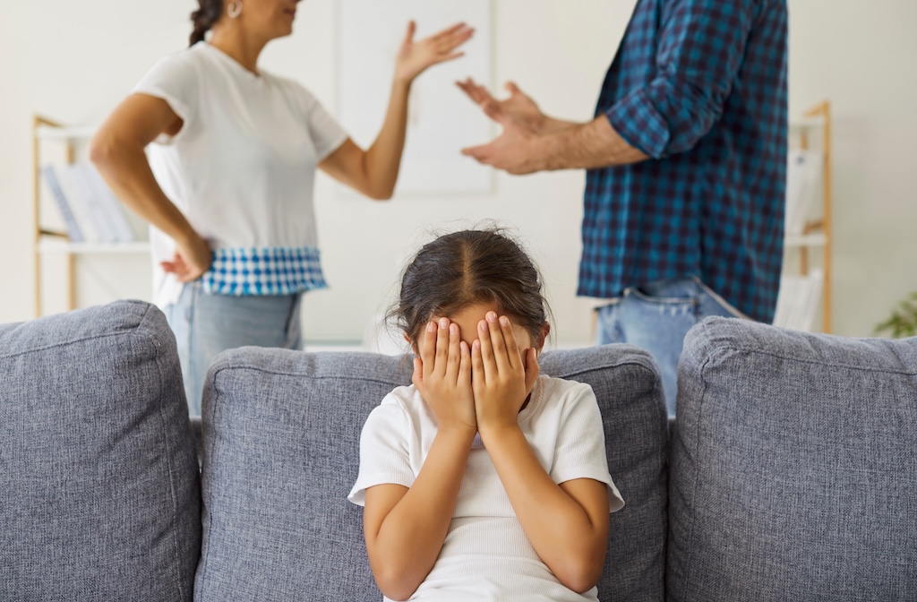 Distressed child covering face as parents argue at home family conflict theme Kids hands hide face amid parental conflict and fight at home. Angry father and mothers arguments and quarrel in living room, sad daughter with stress and trauma crying