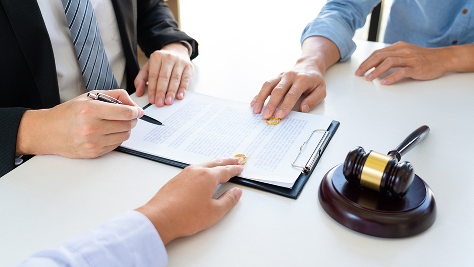 Couple husband and wife during divorce process listening to lawy | Law Office of Eric Andrew Mercer A person in a suit signs a contract on a clipboard while a gavel sits on the desk nearby as others watch.